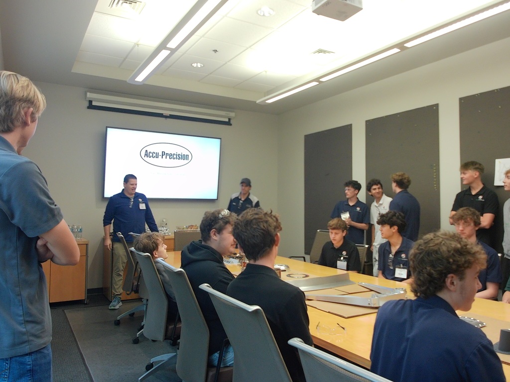 A group of people are seated at tables in a meeting room with a projector screen in the background.