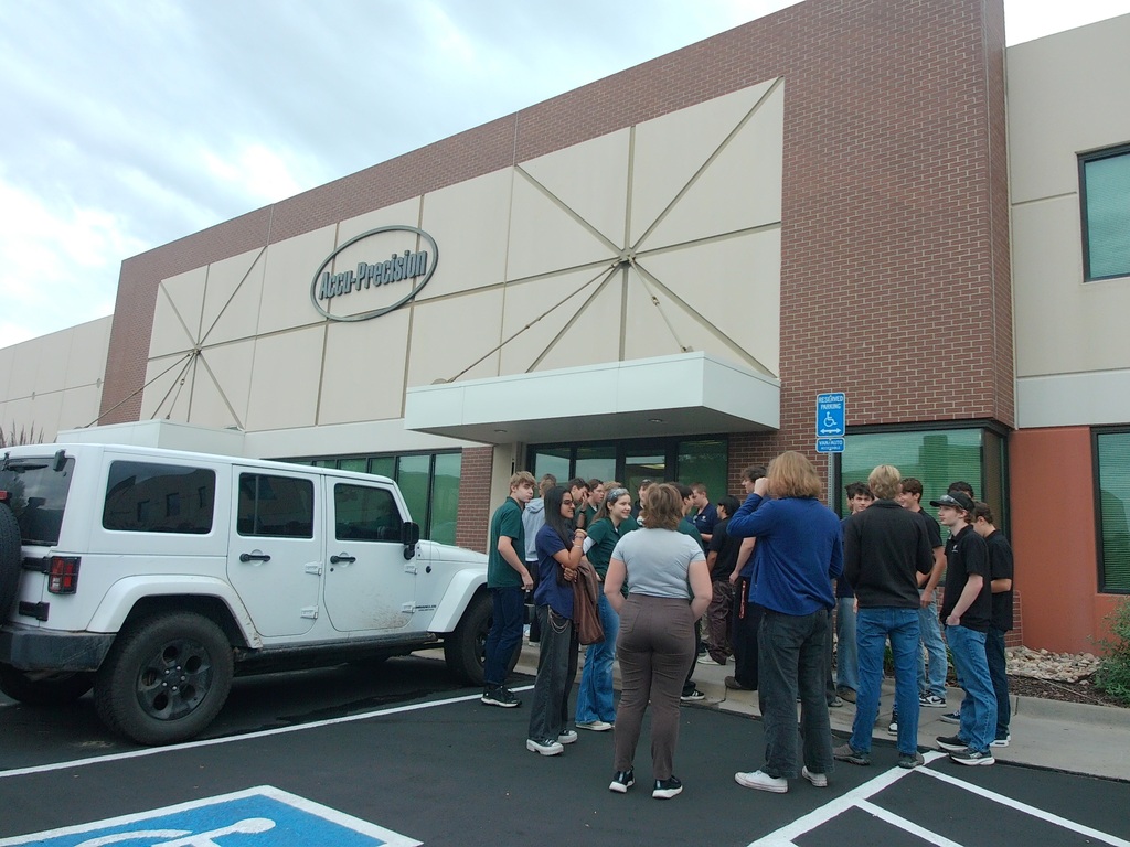 Several people gather in a parking lot. A white Jeep is parked. The building has a logo and large windows.