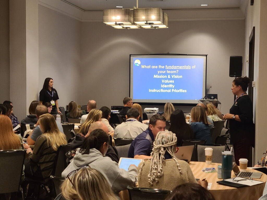 People in a room with a blue screen in the background, listening to a speaker at a conference.