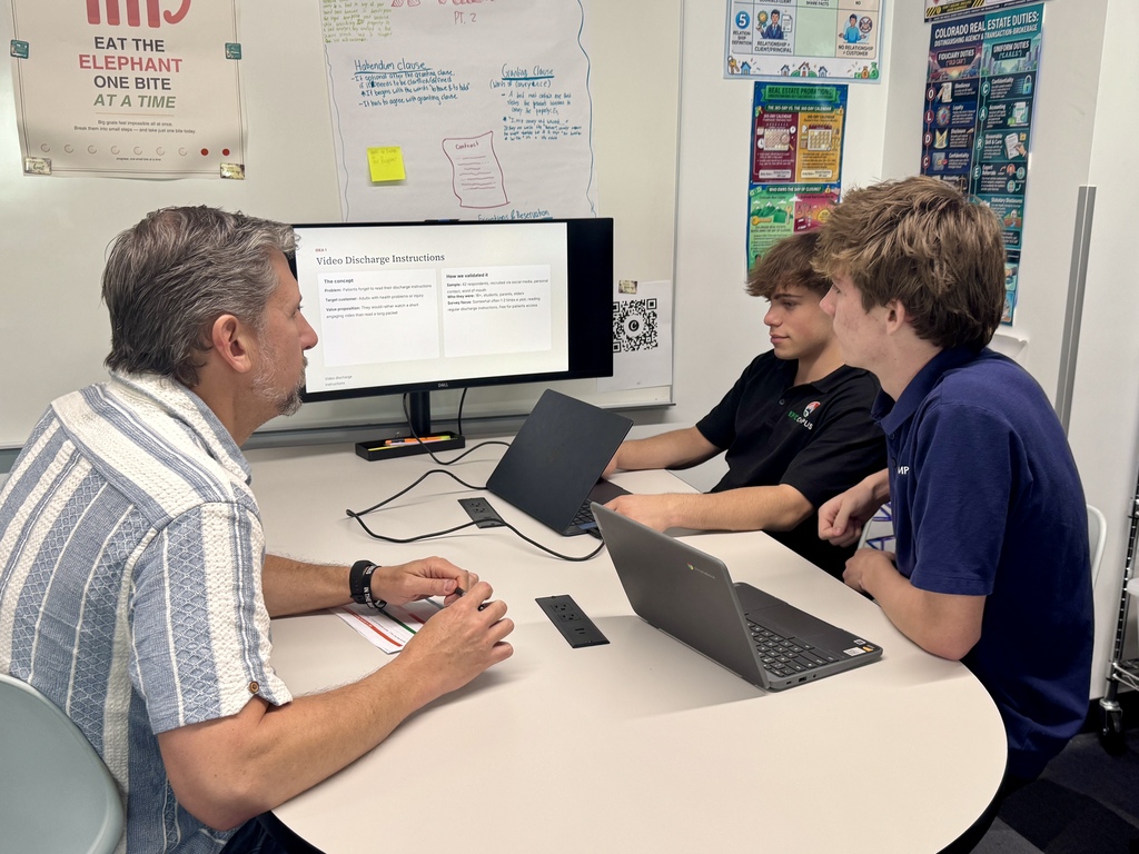 Three people sit around a desk; a man in a striped shirt, two young men in blue shirts. A monitor and laptops are on the desk.