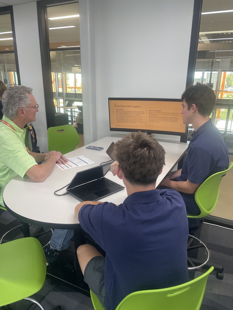 Three people sit around a table. Two men look at a laptop on the table. A man in green wears glasses.