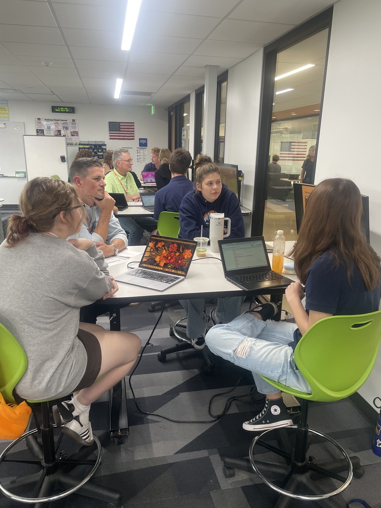 Five people sit around a table with laptops, drinks, and a whiteboard in the background. Chairs are green.