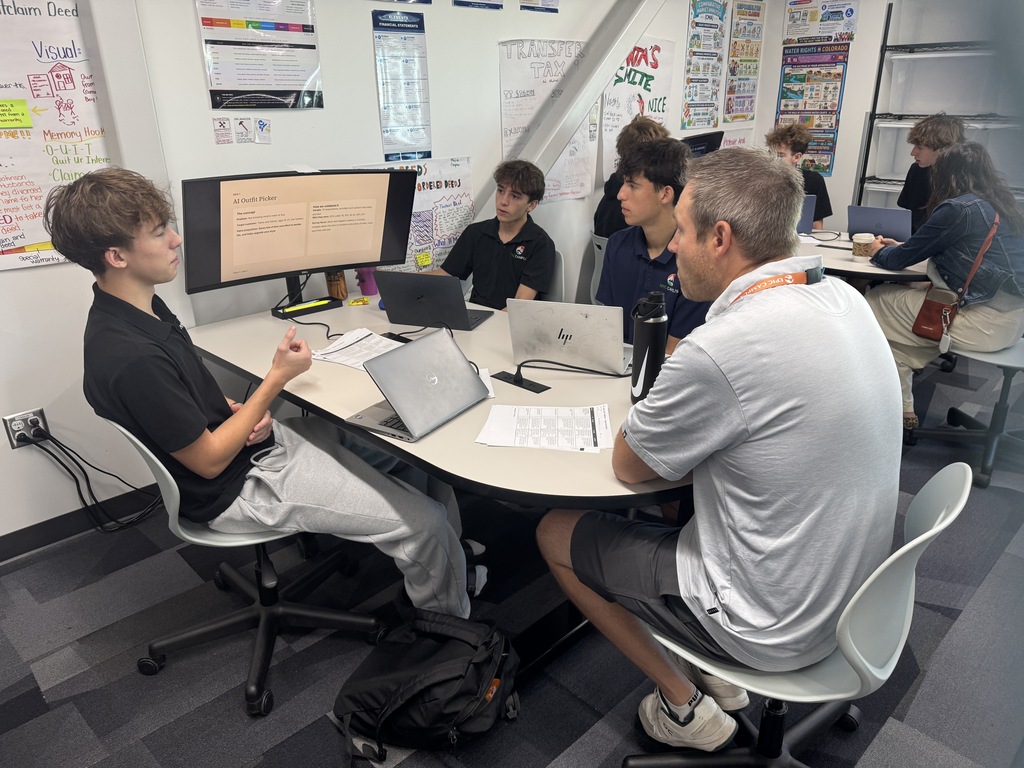 A teacher and student engage in a discussion at a desk with laptops, monitors, and papers.