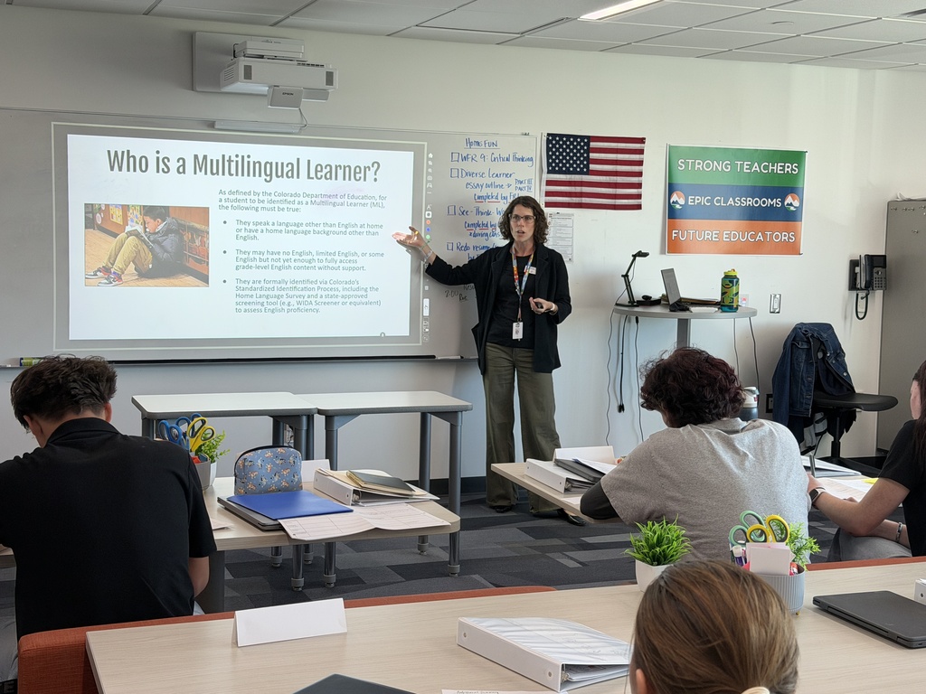 Teacher in front of class. A projector screen shows a question. Students listen. A flag is on the wall.