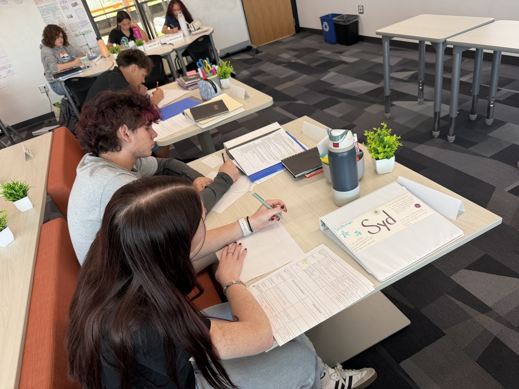 People sit at tables in a room, writing and reading on papers with plants and pens.
