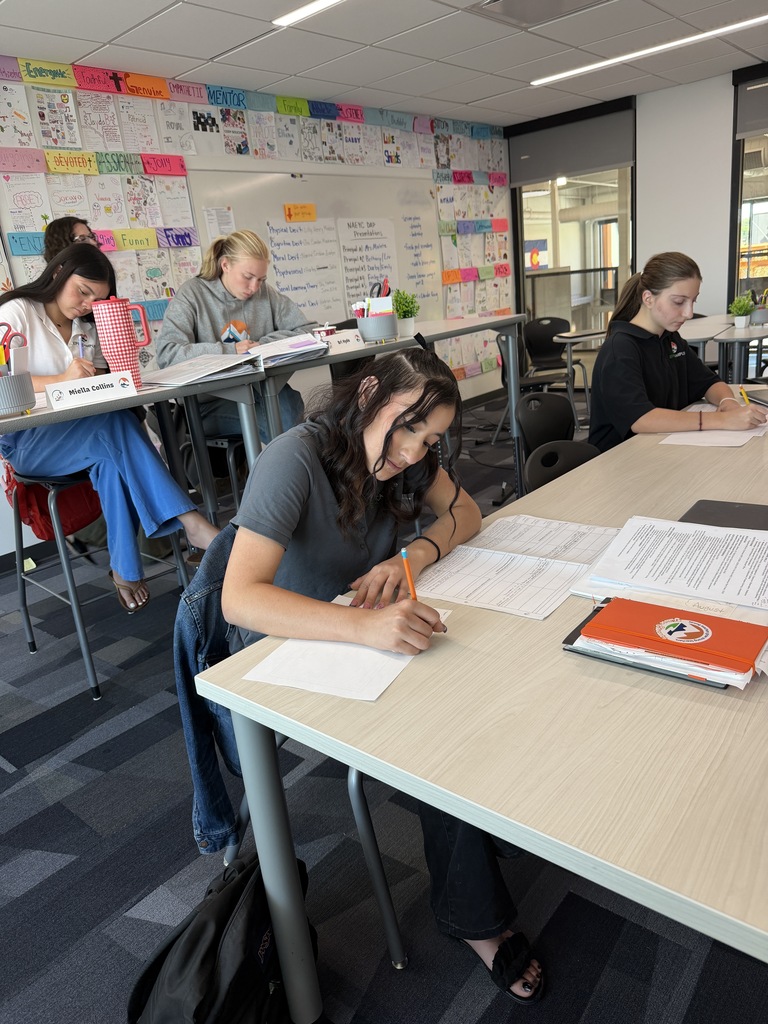 Five students seated at desks in a classroom with a glass window, working on papers and books.