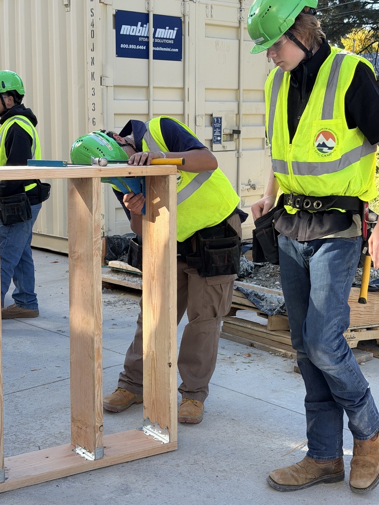 Three workers in helmets and safety vests assemble a wooden structure on a concrete surface.