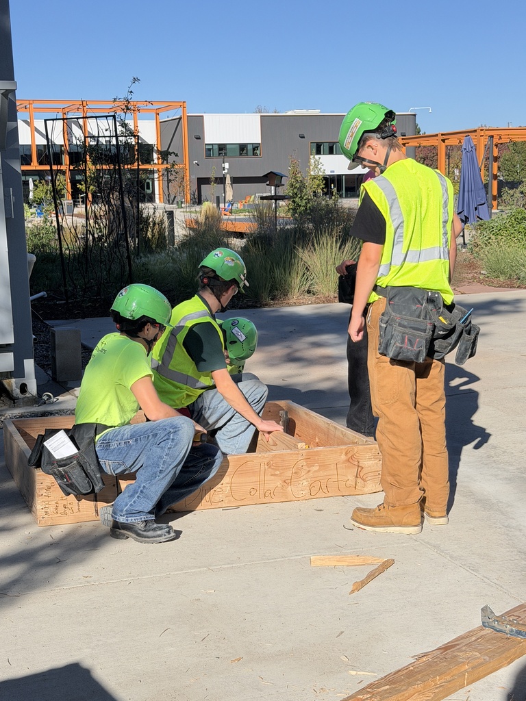 Four workers wearing green helmets and vests work on a raised brick platform outdoors. Behind them, a building is visible.