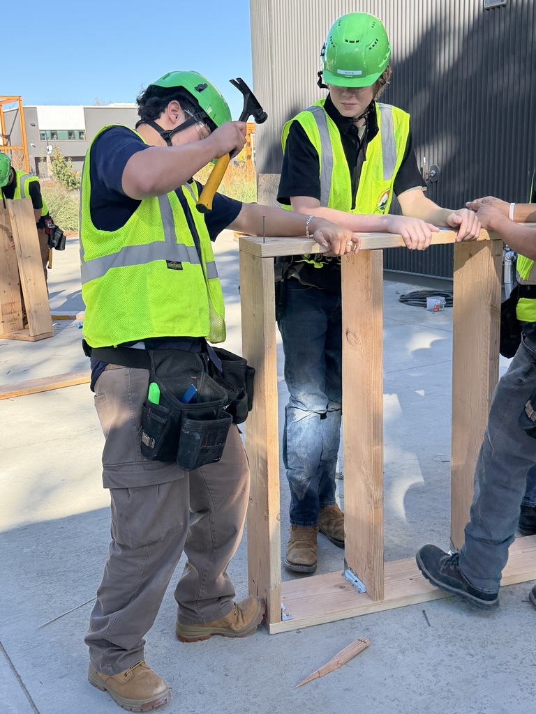 Four workers in safety vests and helmets, constructing a wooden frame with tools. One holds a hammer, while others guide the frame.