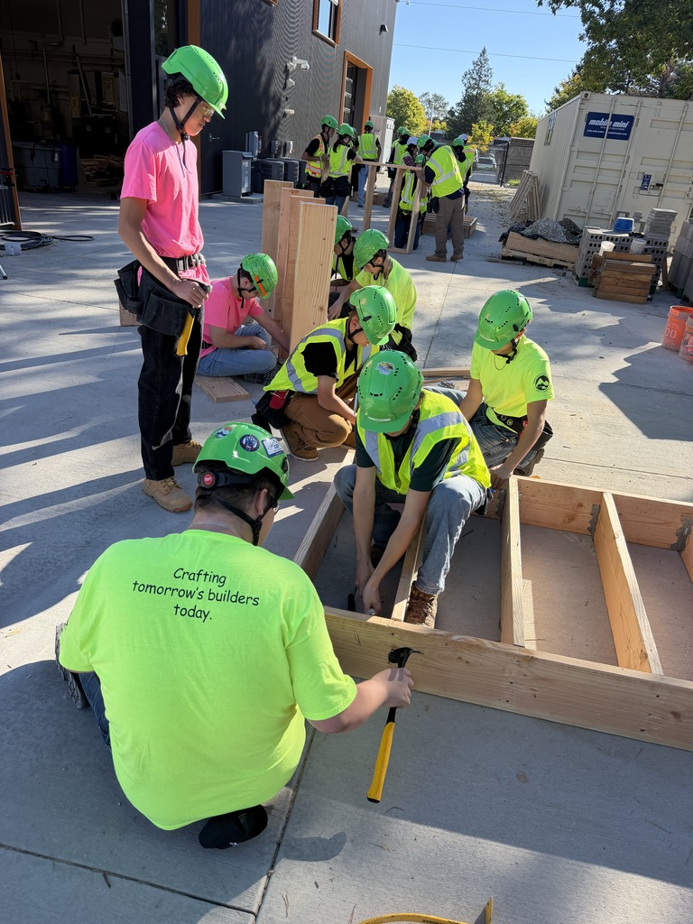 A group of people in yellow vests and helmets work on a wooden structure on a concrete surface.
