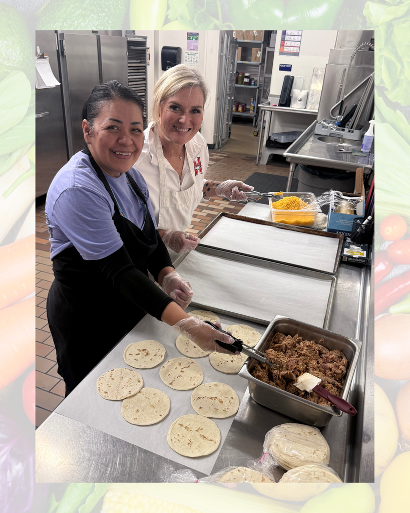 Two women wearing aprons and gloves in a kitchen with trays of tortillas and meat, smiling and posing for a photo.