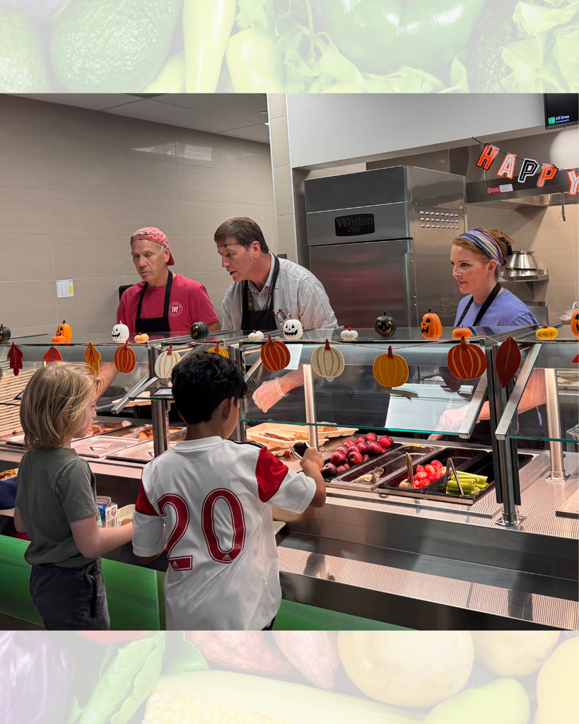 People serve themselves food at a cafeteria. Some pumpkins hang from the serving counter.