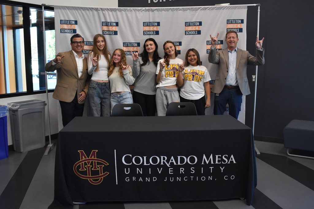 Seven people pose together behind a Colorado Mesa University table, all throwing up hand horns gestures in celebration.