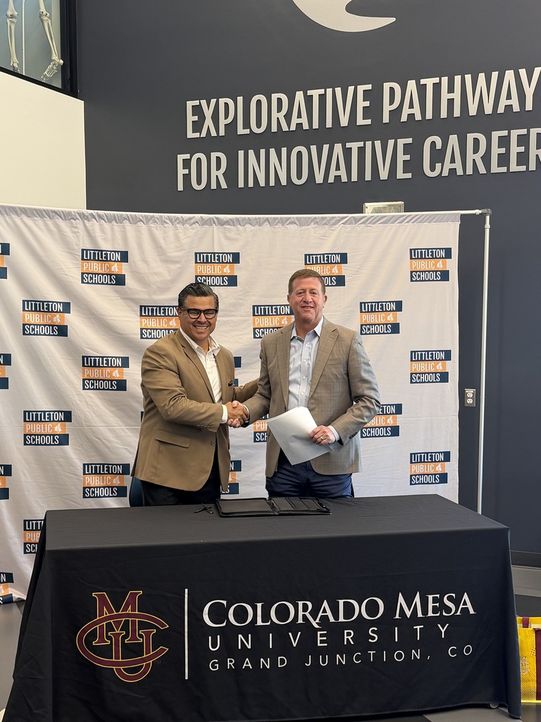 Two men in suits shake hands at a table with a Colorado Mesa University tablecloth and Littleton Public Schools backdrop.