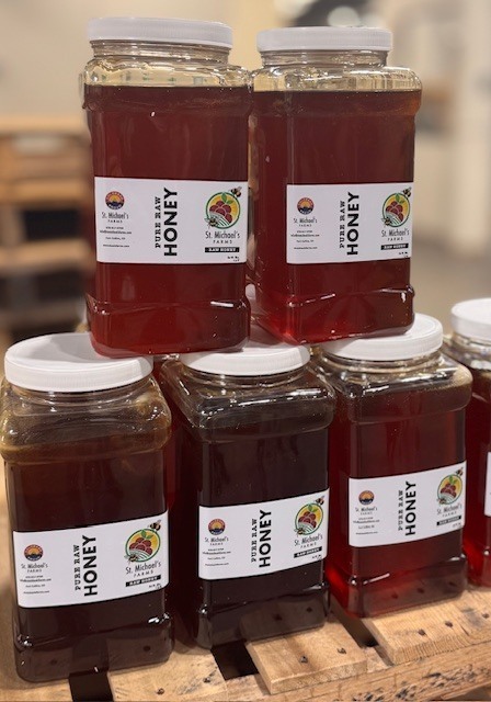 Stacked clear plastic jars of St. Michael’s Farms pure raw honey with white lids sitting on a wooden pallet.