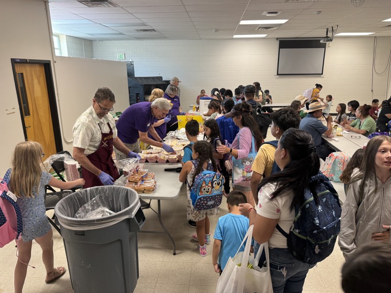 Volunteers in aprons serve food to students and families wearing backpacks lined up in a cafeteria.