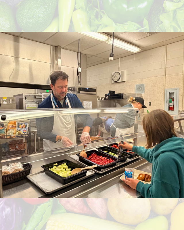 A cafeteria worker serving fresh fruit and vegetables from a salad bar to a student holding a lunch tray.