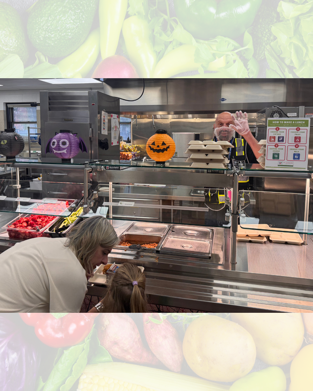 A food service worker waving from behind a glass serving counter with Halloween decorations as a child picks food.