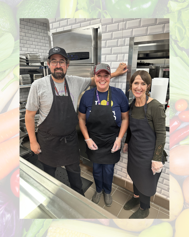 Three school food service staff members wearing aprons and smiling together inside a commercial kitchen area.