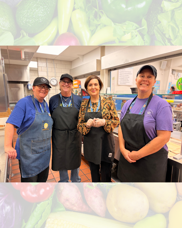 Four cafeteria staff members in aprons posing together behind a serving counter in a school cafeteria.