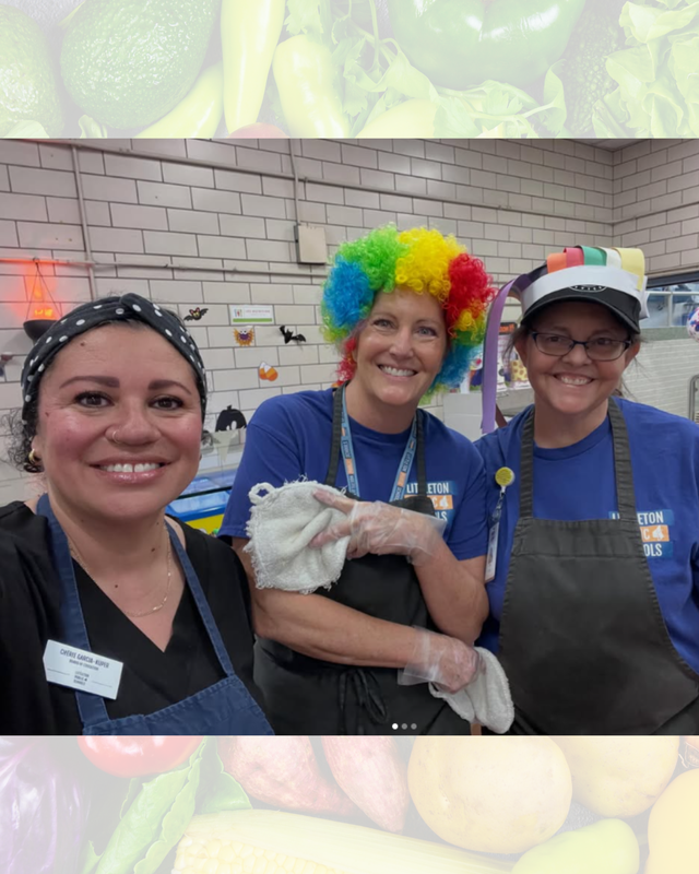 Three school cafeteria staff members smiling together, one wearing a colorful afro wig and another a festive hat.