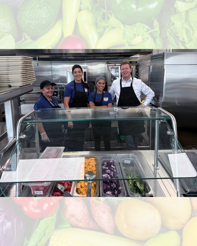 Four cafeteria team members wearing aprons and gloves standing behind a stainless steel serving bar with fresh food.