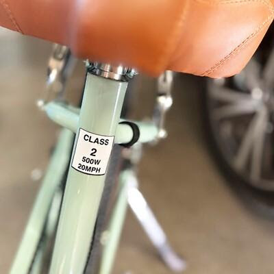 Close-up of a light green e-bike frame under a brown saddle, featuring a label reading Class 2, 500W, 20MPH.