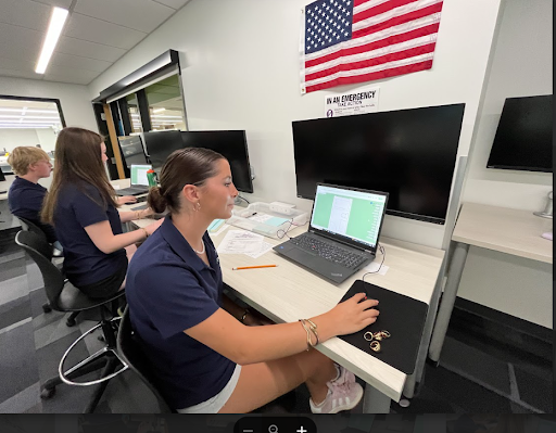  A woman with dark hair, wearing a navy blue polo shirt and white shorts, sits at a desk in an office. She is looking to her right at a computer screen. She has a laptop and a second monitor in front of her, and her right hand is on a computer mouse.
