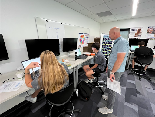 A male teacher stands next to a student, looking at her computer screen at a desk with a laptop and an external monitor. A second student is also working at the same desk, while a third is visible in the background.