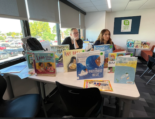  Two people are seated at a table in a classroom with a large display of children's books arranged between them. The woman on the left is looking toward the camera, while the woman on the right is looking away.