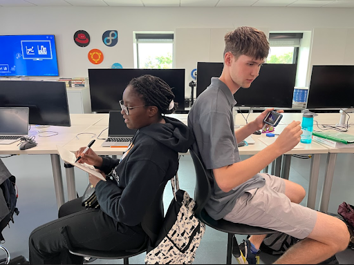  A teenage girl and boy are seated back-to-back in chairs, facing away from each other. The girl is writing on a paper while the boy is looking at his phone.