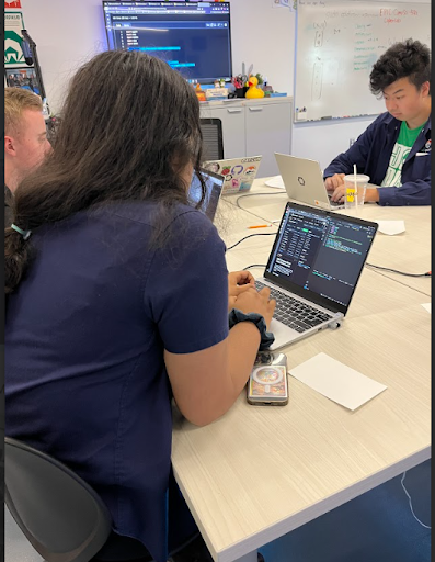  A person with long dark hair is seated at a table, typing on a laptop with code visible on the screen. Two other male students are also at the table, one with blonde hair and the other with black hair, both working on their own laptops.