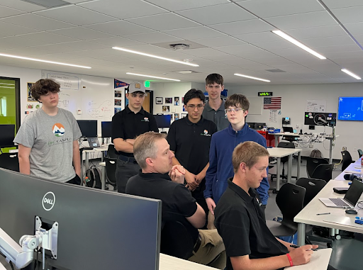 A teacher with short gray hair and a black shirt is seated, facing a group of five male students who are standing around a computer table. The students are looking intently at the computer screen.