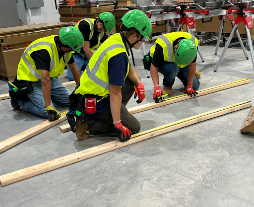  Four male construction students are kneeling on the floor in a workshop, measuring a pair of long wooden boards with tape measures. They are all wearing bright yellow safety vests and green hard hats.