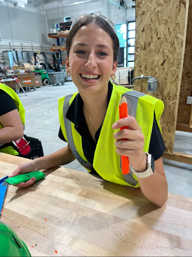  A smiling female student wearing a neon yellow safety vest and goggles is seated at a workbench. She is holding up a large orange pencil in one hand and a utility knife in the other.