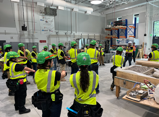  A large group of construction students wearing green hard hats and yellow safety vests are standing in a circle in a workshop. They are doing stretching exercises together, with their arms extended in front of them.