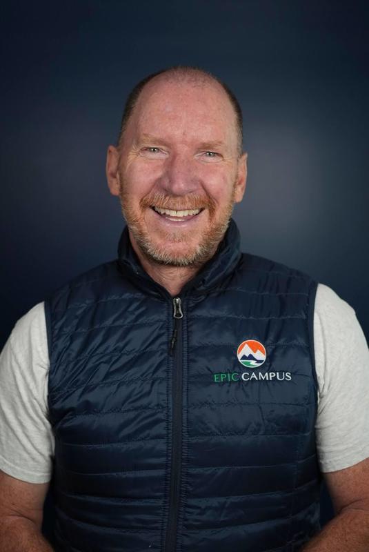  A smiling, middle-aged man with a reddish beard and a bald head is wearing a navy blue puffer vest over a white shirt. The vest has a logo on the left side that reads "EPIC CAMPUS" with a small circular graphic above it.