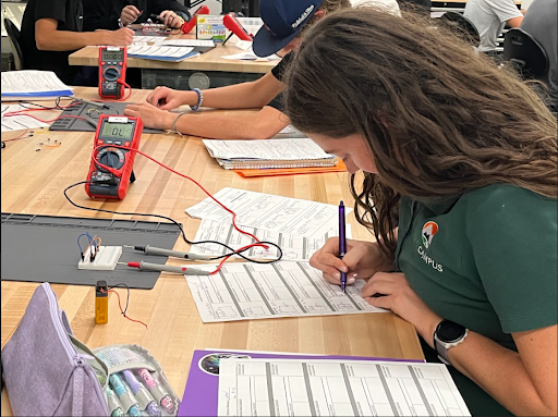 A person with long, brown hair is writing on a worksheet at a desk with a purple pen. A red digital multimeter and a small electronic circuit are visible on the desk. Other students are working with similar equipment nearby.