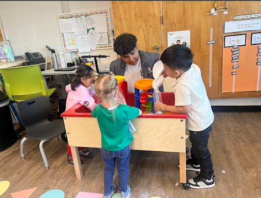 A young man and three children are playing at a wooden sand and water table in a classroom. The man is helping a young boy pour water into a colorful plastic water funnel set.