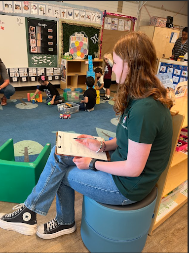 A young woman is seated on a low stool, writing on a clipboard. She is wearing a dark green polo shirt, blue jeans, and black Converse shoes. Several young children are playing in the background.