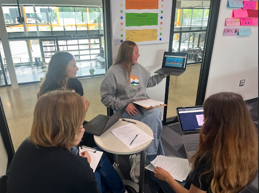 A group of four young women is seated in a semicircle in a modern, open-concept office space. One woman is holding up a laptop to show the others something on the screen, while the rest are looking on with papers and clipboards in hand.