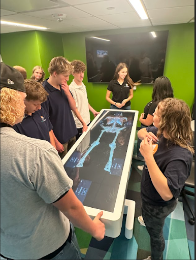 A group of high school students is gathered around a large interactive digital anatomy table, which displays a glowing human skeleton. A female instructor is explaining the display to the students.