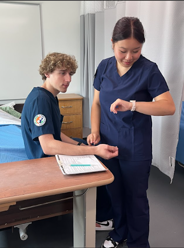 A young man and woman, both dressed in navy blue scrubs, are in a simulated patient room. The woman is checking a watch on her wrist as she takes the pulse of the young man, who is seated on a bed.
