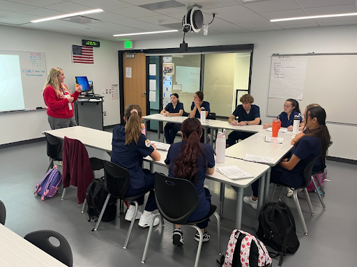 A teacher in a red hoodie is standing in front of a classroom, gesturing with her hands while speaking to a group of seven students. The students are seated around two white tables pushed together, wearing navy blue scrubs.