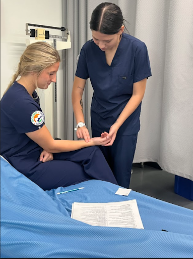 Two young women, both wearing navy blue scrubs, are in a simulated patient room. One woman is seated on a bed while the other stands beside her, taking her pulse.