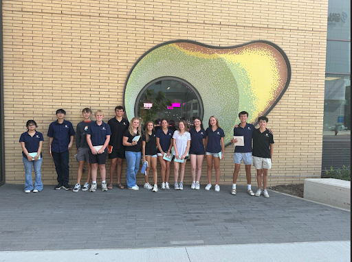  A group of high school students is posing for a photo in front of a modern building with a distinctive, gourd-shaped mosaic window. Most of the students are wearing dark blue polo shirts and holding notebooks or papers.