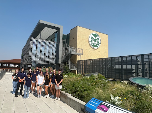 A group of high school students is posing for a photo on a sunny day in front of a modern building with a Colorado State University Rams logo on it. The building has a prominent green roof with lush vegetation and large windows.
