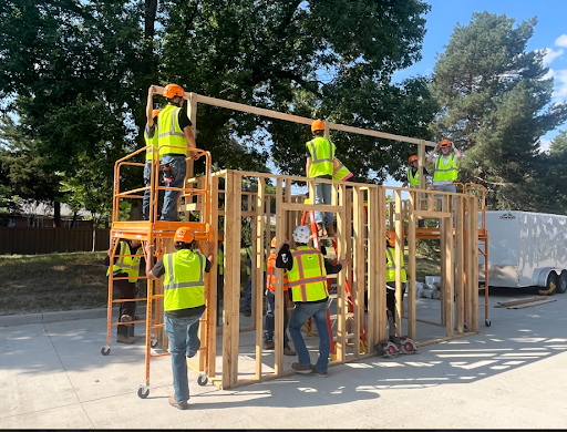  A group of people wearing safety vests and hard hats work together to build a wooden frame structure outdoors, using ladders and scaffolding.