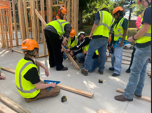  A group of young people wearing safety vests and hard hats are gathered around a piece of lumber on the ground, actively working on a construction project.