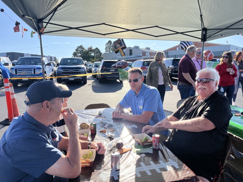 Three men sit at a table under a tent while talking and eating at a tailgate party.